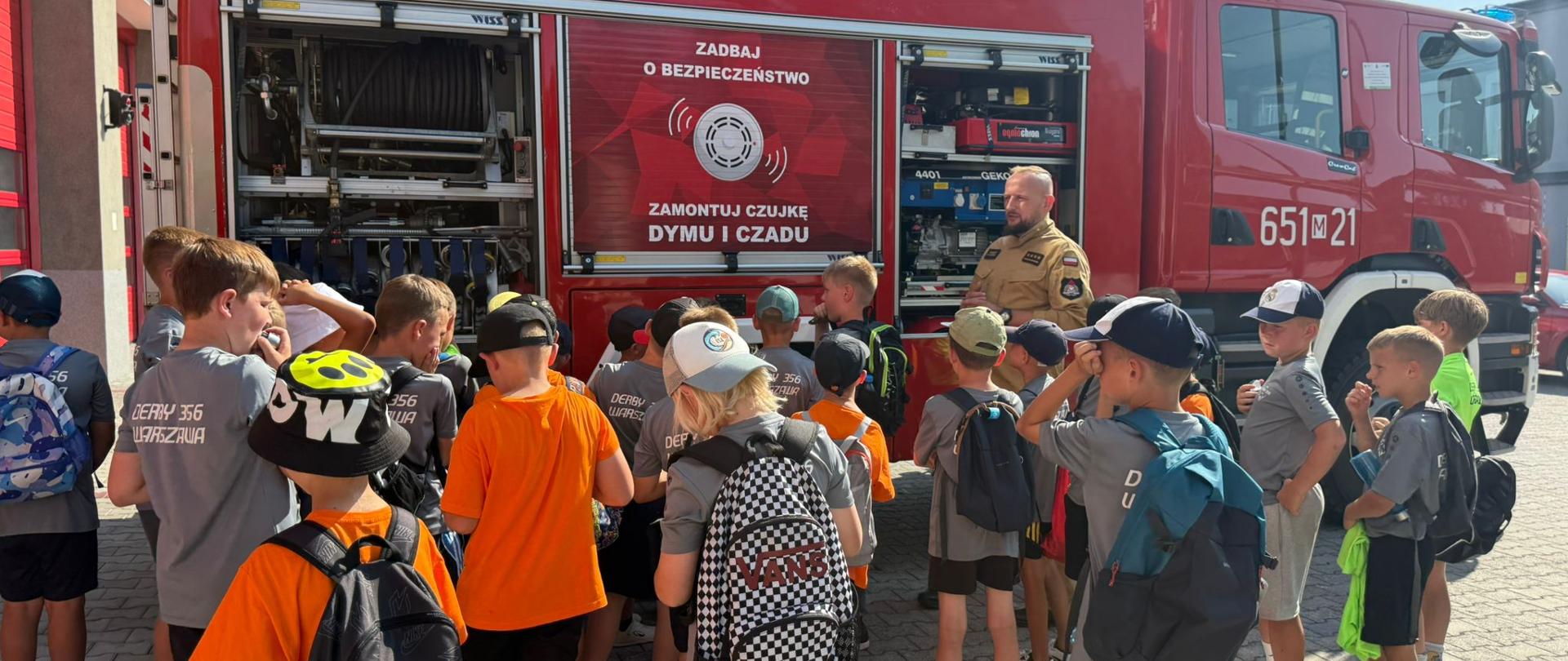 Zdjęcie przedstawia grupkę dzieci, które słuchają strażaka opowiadającego im o sprzęcie znajdującym się na wyposażeniu JRG. Za strażakiem duży czerwony samochód z naklejką na której jest napis: zadbaj o bezpieczeństwo, zamontuj czujkę dymu i czadu. 