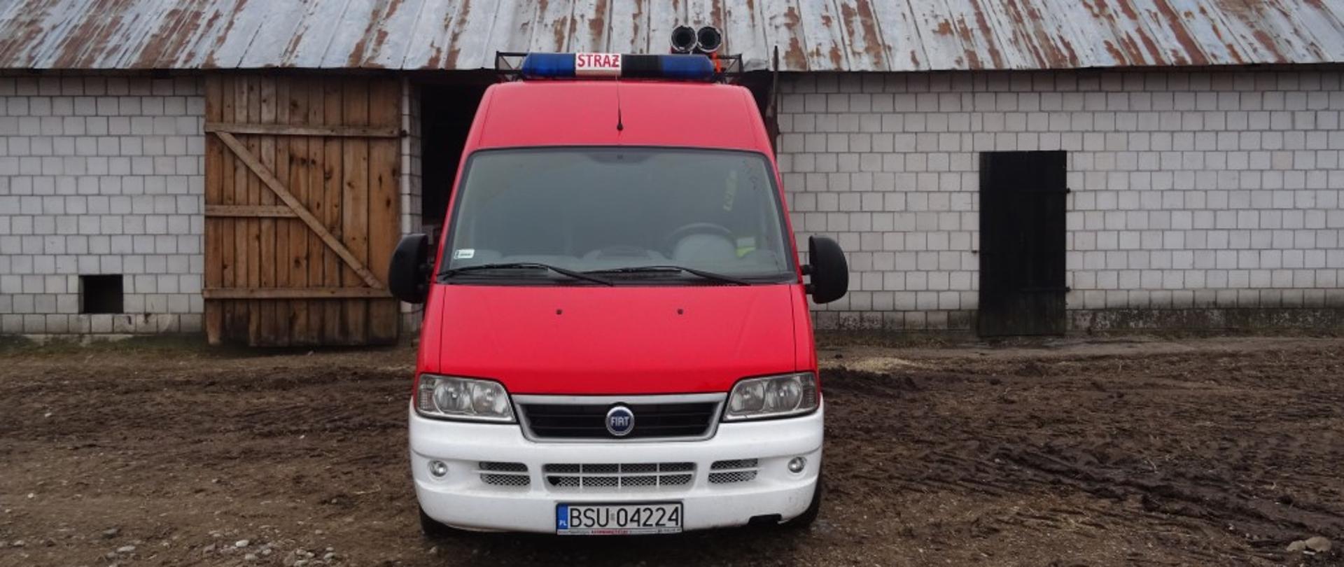 Na pierwszym planie pojazd ratowniczy koloru czerwonego z białym zderzakiem. W tle budynek gospodarczy do przechowywania auta.