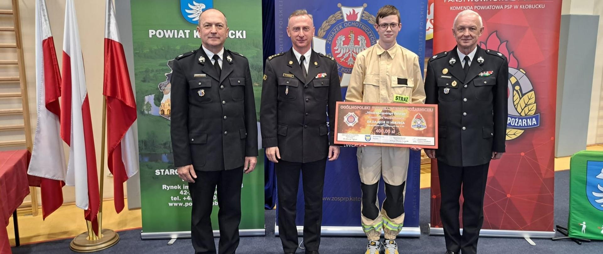 Cztery osoby stoją do zdjęcia w sali gimnastycznej na tle banerów instytucji strażackich i samorządowych oraz flag Polski. Trzech mężczyzn ubranych jest w strażackie mundury galowe , a między nimi stoi młody chłopak w jasnym mundurze Młodzieżowej Drużyny Pożarniczej, trzymający dyplom za zajęcie 3. miejsca. Chłopak pochodzi z powiatu kłobuckiego, druh młodzieżowej drużyny pożarniczej OSP Zajączki Drugie. Wśród osób dorosłych znajdują się wiceminister edukacji Henryk Kiepura, Śląski Komendant Wojewódzki PSP st. bryg. Damian Legierski oraz starosta kłobucki Dariusz Pilśniak. 