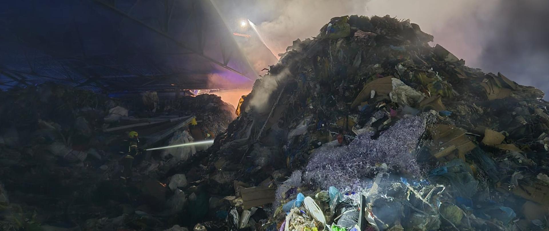 Zdjęcie przedstawia palącą się pryzmę odpadów. Z lewej strony stoi strażak podając wodę na palącą się pryzmę.