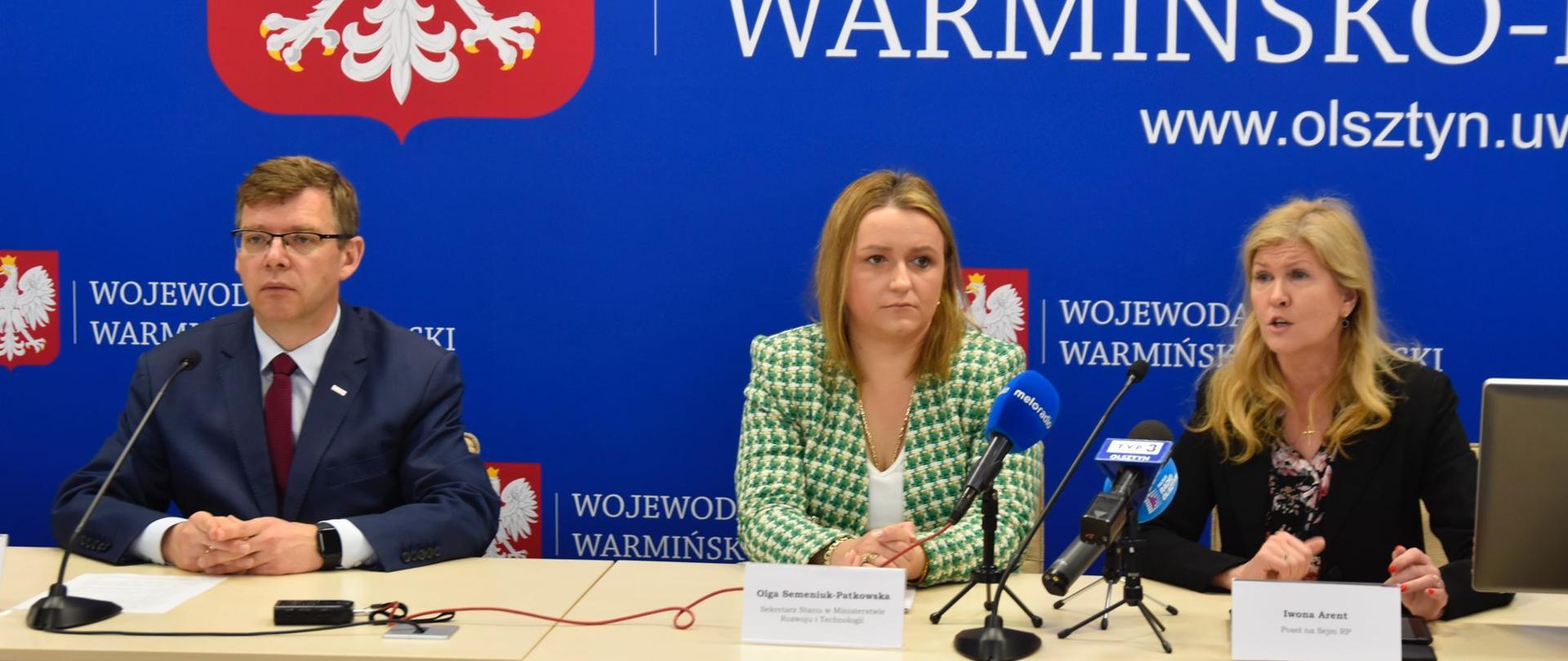 Wojewoda oraz dwie kobiety siedzą przy stole z mikrofonami i przemawiają