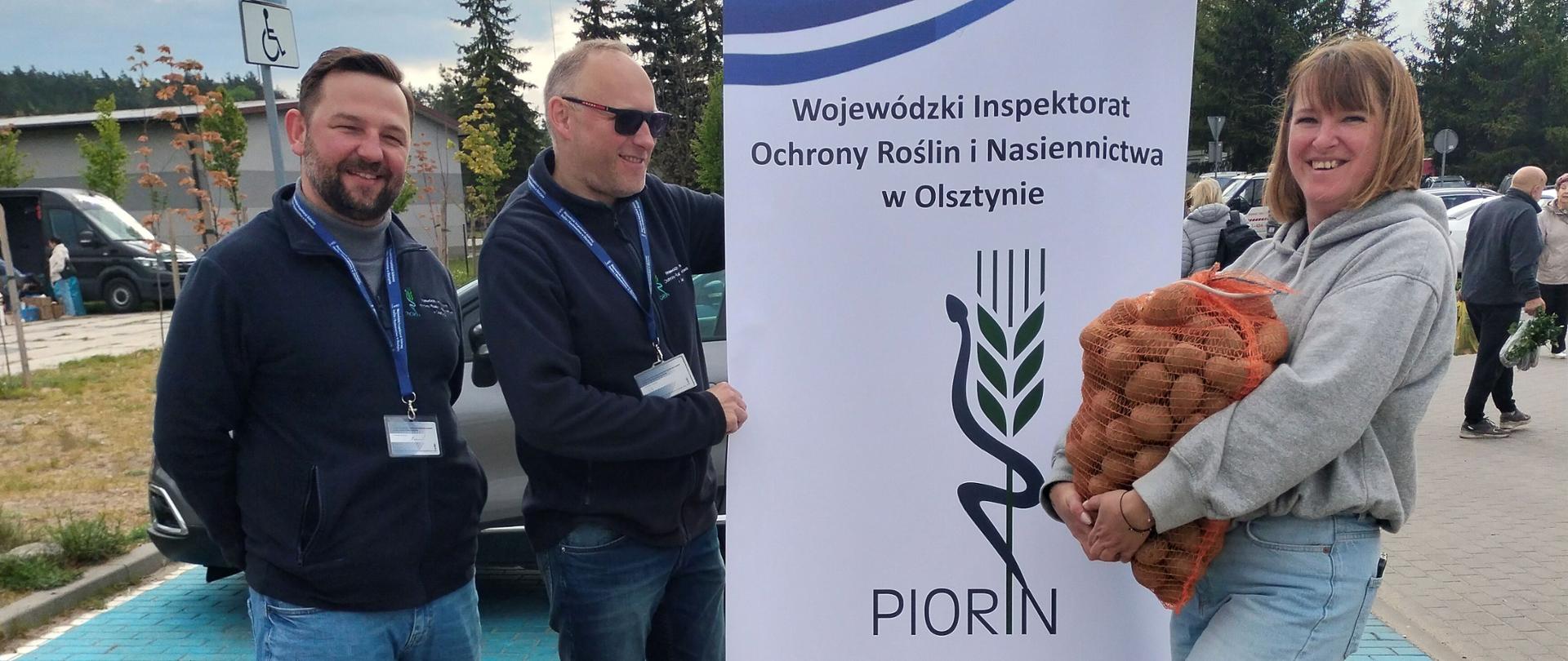  Trzy uśmiechnięte osoby przy plenerowym stoisku informacyjnym. Dwóch mężczyzn stoi obok stolika z ulotkami, a kobieta trzyma duży worek ziemniaków obok firmowego baneru