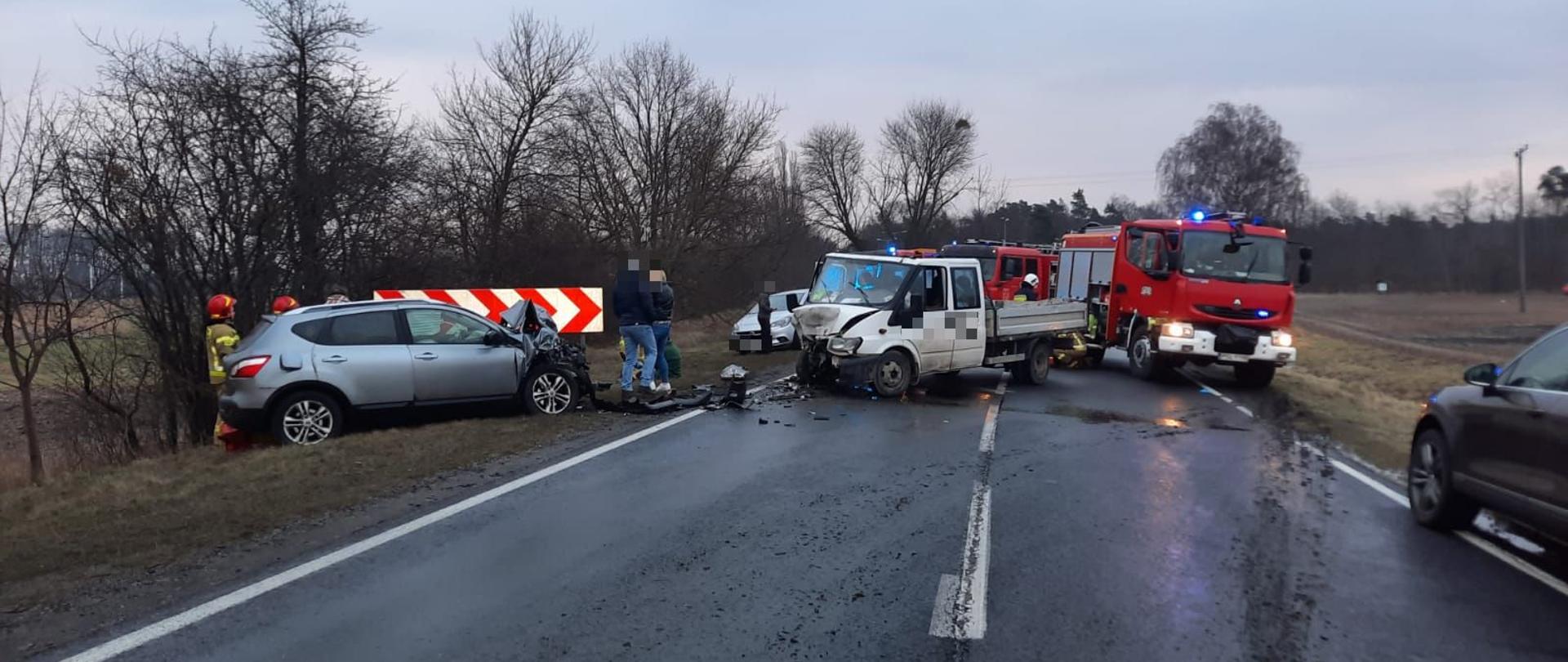 rozbite samochody srebrny osobowy oraz biały typu bus za srebrnym samochodem stoją strażacy przed samochodem dwie osoby postronne za busem stoją dwa samochody straży pożarnej, za srebrnym samochodem krzaki oraz drzewa 