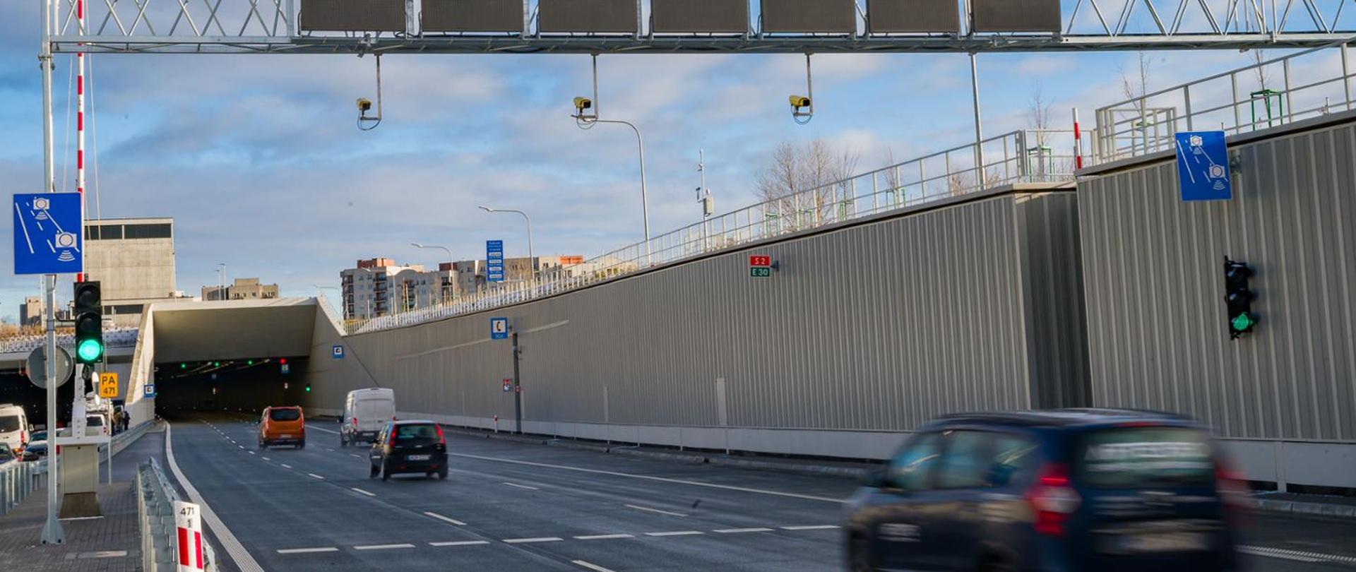 Wjazd do tunelu na Ursynowie, trzypasowa trasa S2, po obu stronach drogi znaki informujące o odcinkowym pomiarze prędkości, w oddali bloki