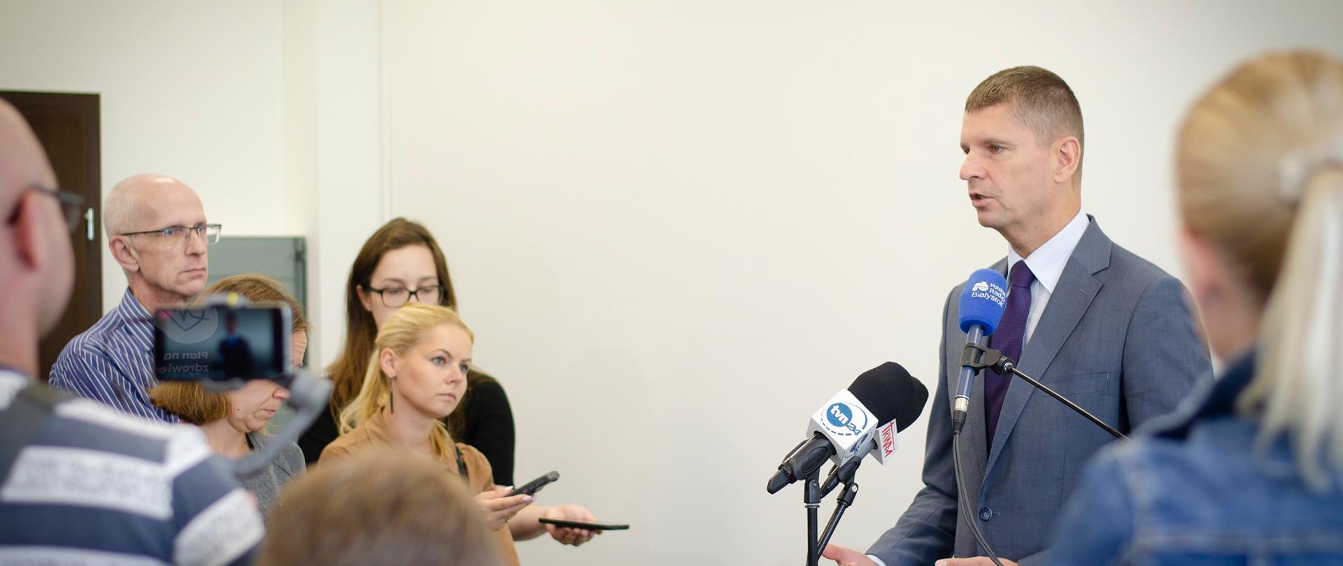 Minister stoi na tle białej ściany, przed nim dwa mikrofony na stojakach i grupa ludzi.