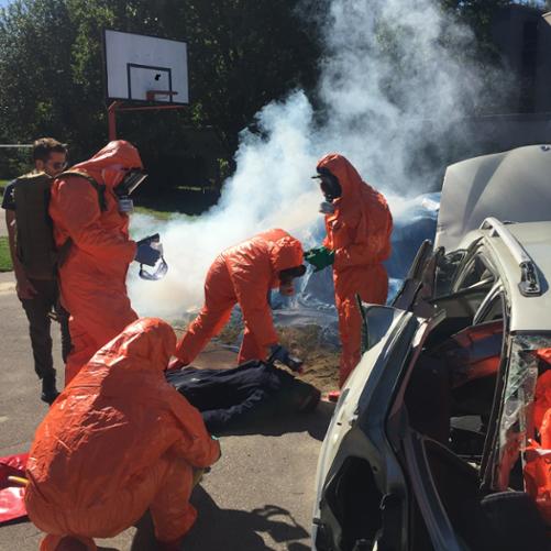 Czterech uczestników szkolenia w ochronnych kombinezonach i maskach sprawdza stan ofiary wypadku leżącej obok rozbitego samochodu
