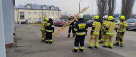 Strażacy Ochotniczej Straży Pożarnej przyglądają się innym strażakom którzy trzymają drabinę, w tle za nimi stoją samochody i widać w oddali budynek koloru beżowego