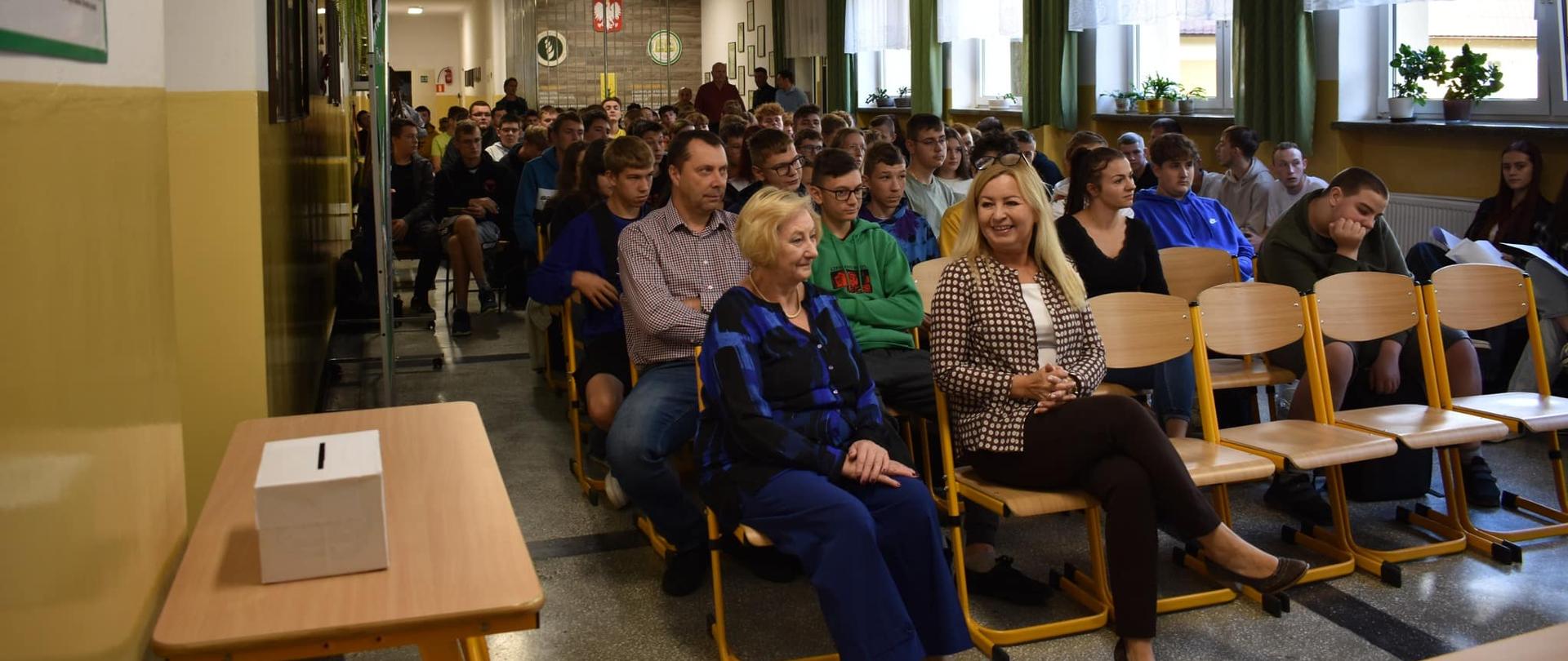 Szerokie ujęcie sali wypełnionej siedzącymi uczniami i nauczycielami. Na pierwszym planie po lewej stronie znajduje się stolik z białą urną wyborczą.