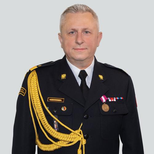 Portretowe zdjęcie funkcjonariusza Państwowej Straży Pożarnej w czarnym mundurze galowym, wykonane na jasnym, jednolitym tle. Mężczyzna stoi przodem do obiektywu, patrzy wprost, ma krótkie siwe włosy i spokojny wyraz twarzy. Na mundurze widoczne są złote dystynkcje, odznaki, baretkowe odznaczenia, medal oraz złoty sznur galowy przewieszony przez prawe ramię i pierś. Na lewej piersi znajduje się imienna plakietka z nazwiskiem „Sierpatowski”.