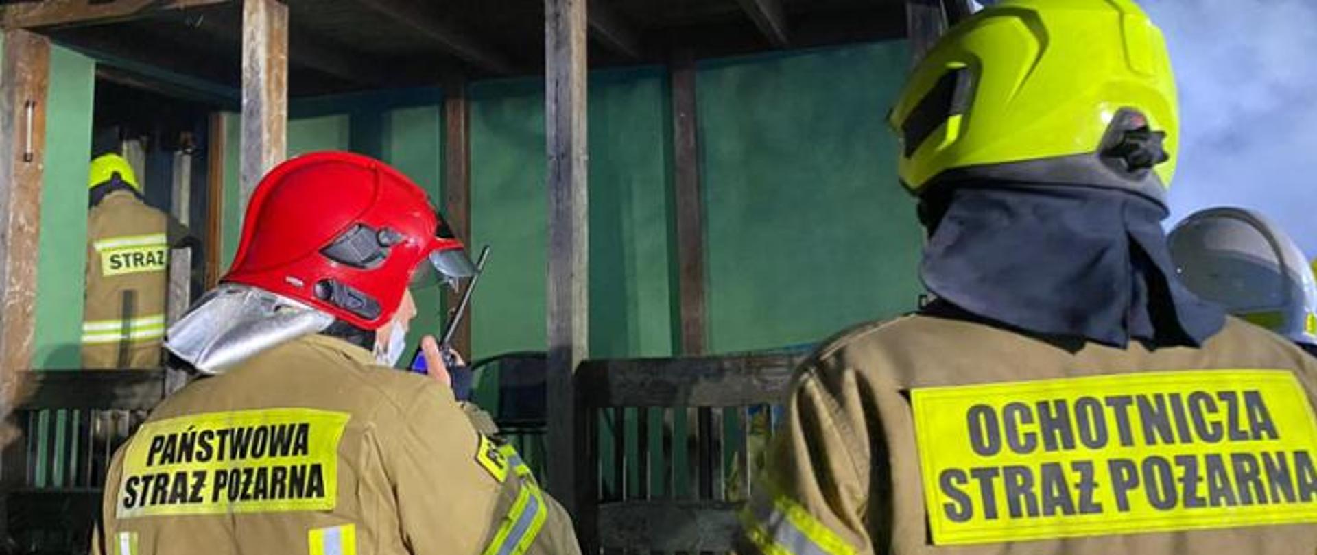Zdjęcie przedstawia kilku strażaków Państwowej i Ochotniczej Straży Pożarnej przed budynkiem podczas akcji gaśniczej. Strażak Państwowej Straży Pożarnej rozmawia przez radiostację przenośną.