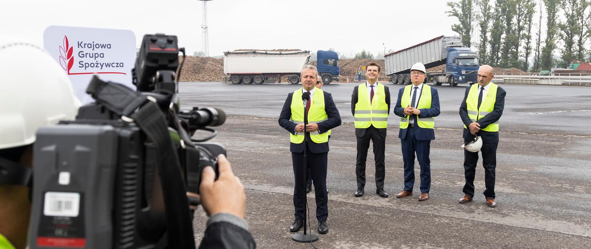 Jeden mężczyzna stojący przed mikrofonem i wypowiadający się. Za nim trzech mężczyzn 