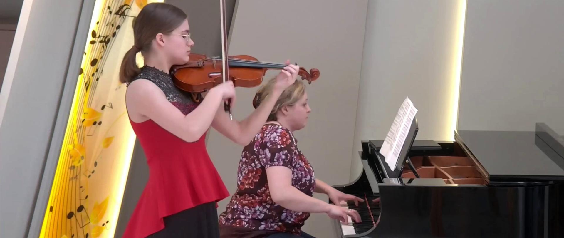 Zdjęcie - koncert w wykonaniu skrzypaczki i towarzyszącej jej pianistki, grającej na fortepianie. W tle podświetlona dekoracja estrady sali kameralnej.
