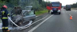 Zdjęcie przedstawia strażaka, który trzymając w ręku prądownice podaje środek gaśniczy na palący się samochód. W tle droga ekspresowa na której stoi samochód pożarniczy oraz poruszające się tą drogą samochody