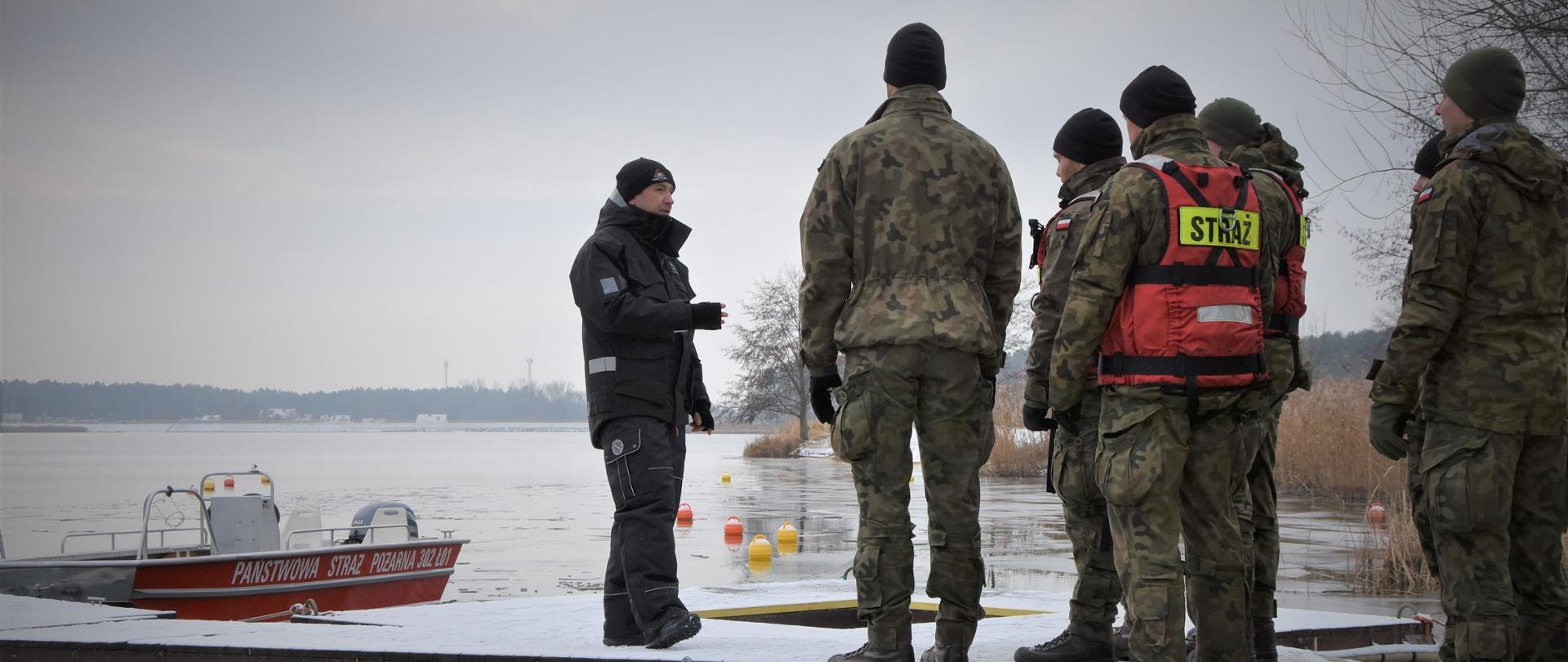 Zdjęcie przedstawia żołnierzy oraz strażaka z JRG 2 PSP w Kielcach. Stoją na pomoście. Za nimi widać łódź Państwowej Straży Pożarnej.