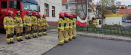 Zdjęcie przedstawia strażaków w Jednostki Ratowniczo-Gaśniczej PSP w Lwówku Śląskim podczas uroczystej zmiany służby w trakcie podniesienia Flagi Państwowej z okazji rocznicy odzyskania niepodległości przez Polskę.
