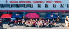 Szeroki widok z góry na elewację budynku Komendy Wojewódzkiej PSP w Opolu. Biały budynek z czerwonymi akcentami i rzędami okien. Na górze duży czerwony napis: „KOMENDA WOJEWÓDZKA PAŃSTWOWEJ STRAŻY POŻARNEJ”. Przed budynkiem, na placu, stoi bardzo duża grupa ludzi – kilkaset osób – ustawionych do wspólnego zdjęcia. Wszyscy w różnych mundurach strażackich (dorośli i dzieci, w tym w mundurach młodzieżowych). Przed grupą, pośrodku, widać dwa czerwone namioty. Lewy namiot z numerem alarmowym 112, prawy z napisem na niebieskim banerze. Plac jest pusty przed grupą, widać tylko cienie. W tle u góry, na dachu, widać instalację fotowoltaiczną.