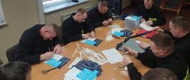 Strażacy w świetlicy na dużym stole naklejają na otrzymane ulotki dane kontaktowe punktów konsultacyjnych dotyczących szczepień. Łącznie 8 osób pracuje nad przygotowaniem ulotek informacyjnych.