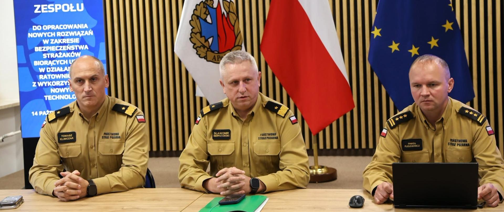 Zdjęcie 6: Trzech funkcjonariuszy Państwowej Straży Pożarnej siedzi za stołem podczas spotkania zespołu. Za nimi znajdują się flagi Akademii Pożarniczej, Polski i Unii Europejskiej oraz ekran z informacją o spotkaniu.