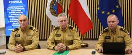 Zdjęcie 6: Trzech funkcjonariuszy Państwowej Straży Pożarnej siedzi za stołem podczas spotkania zespołu. Za nimi znajdują się flagi Akademii Pożarniczej, Polski i Unii Europejskiej oraz ekran z informacją o spotkaniu.