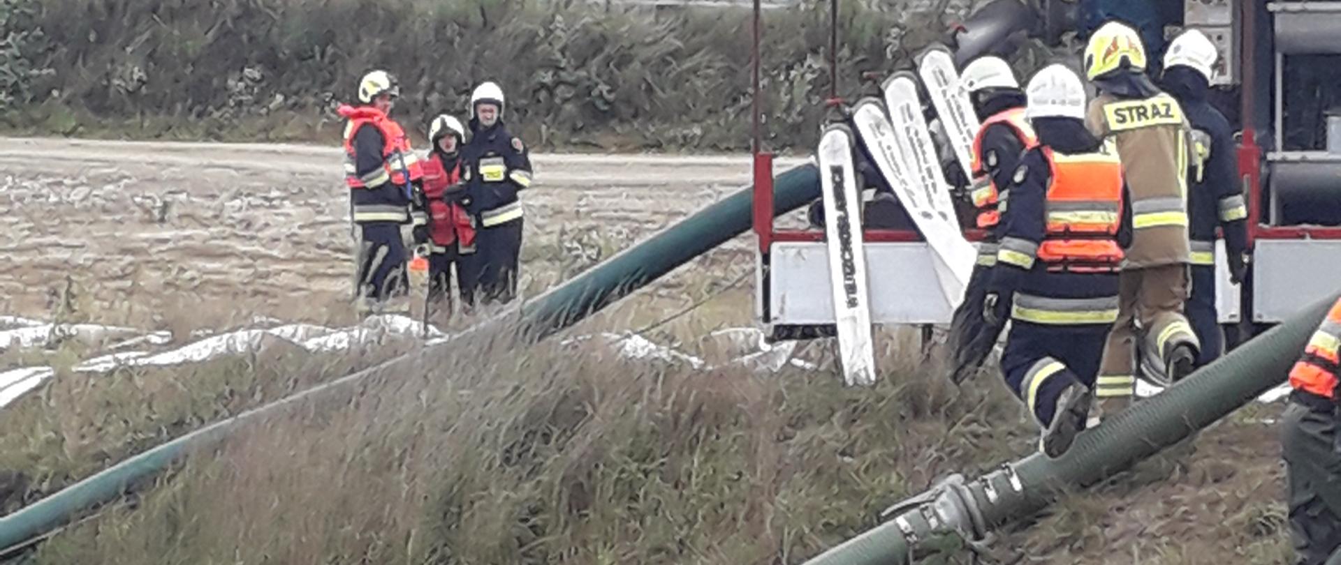 Na zdjęciu widoczni są strażacy, którzy pracują przy rozstawianiu pompy wysokiej wydajności. Węże ssawne wrzucone są do zbiornika wodnego a strażacy rozkładają węże tłoczne. 