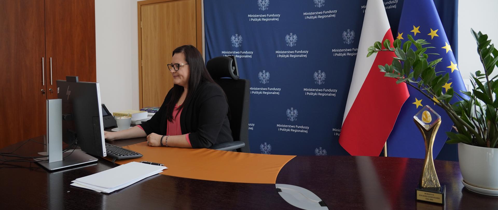 Wiceminister Małgorzata Jarosińska-Jedynak siedzi w gabinecie przy biurku z monitorem. Za nią ścianka MFiPR i flagi PL i UE. Obok kwiaty w doniczce.