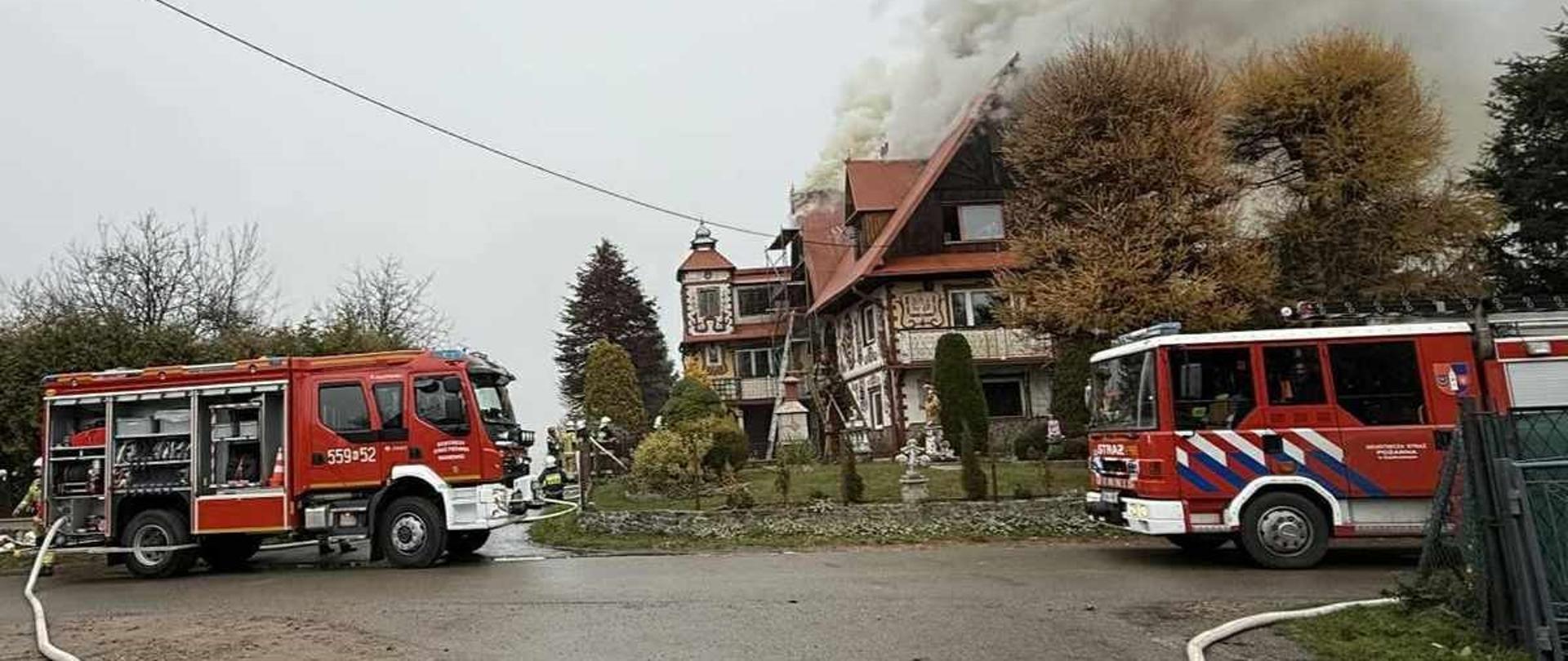 Na zdjęciu widać dwa samochody pożarnicze stojące przed budynkiem, z którego wydobywa się gęsty dym. Na asfalcie leżą rozwinięte linie wężowe a strażacy prowadzą działania związane z gaszeniem pożaru poddasza.