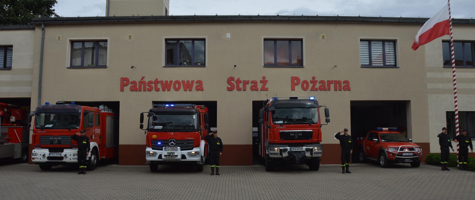79 rocznica wybuchu Powstania Warszawskiego. Na tle budynku komendy powiatowej PSP stoi 5 pojazdów pożarniczych z włączoną sygnalizacją świetlną. Obok każdego z nich stoi strażak w ubraniu koszarowym, który oddaje honor poprzez salutowanie. Z prawej strony na maszcie powiewa flaga biało-czerwona