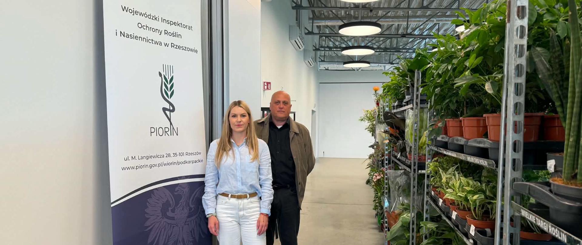 Na zdjęciu widać Inspektorów WIORiN w Ustrzykach Dolnych stojących w nowoczesnym wnętrzu FANTO Bieszczadzkiego Centrum Dziedzictwa Kulturowego. Po lewej stronie znajduje się roll-up z logo PIORiN i napisem Wojewódzkiego Inspektoratu Ochrony Roślin i Nasiennictwa w Rzeszowie. Kobieta ubrana jest w jasnoniebieską koszulę i białe spodnie, obok niej stoi mężczyzna w ciemnym ubraniu i brązowej kurtce. Po prawej stronie ustawione są metalowe regały z licznymi roślinami doniczkowymi. W tle widoczne są wysokie sufity z industrialnym wykończeniem i okrągłe lampy sufitowe.
