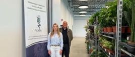 Na zdjęciu widać Inspektorów WIORiN w Ustrzykach Dolnych stojących w nowoczesnym wnętrzu FANTO Bieszczadzkiego Centrum Dziedzictwa Kulturowego. Po lewej stronie znajduje się roll-up z logo PIORiN i napisem Wojewódzkiego Inspektoratu Ochrony Roślin i Nasiennictwa w Rzeszowie. Kobieta ubrana jest w jasnoniebieską koszulę i białe spodnie, obok niej stoi mężczyzna w ciemnym ubraniu i brązowej kurtce. Po prawej stronie ustawione są metalowe regały z licznymi roślinami doniczkowymi. W tle widoczne są wysokie sufity z industrialnym wykończeniem i okrągłe lampy sufitowe.