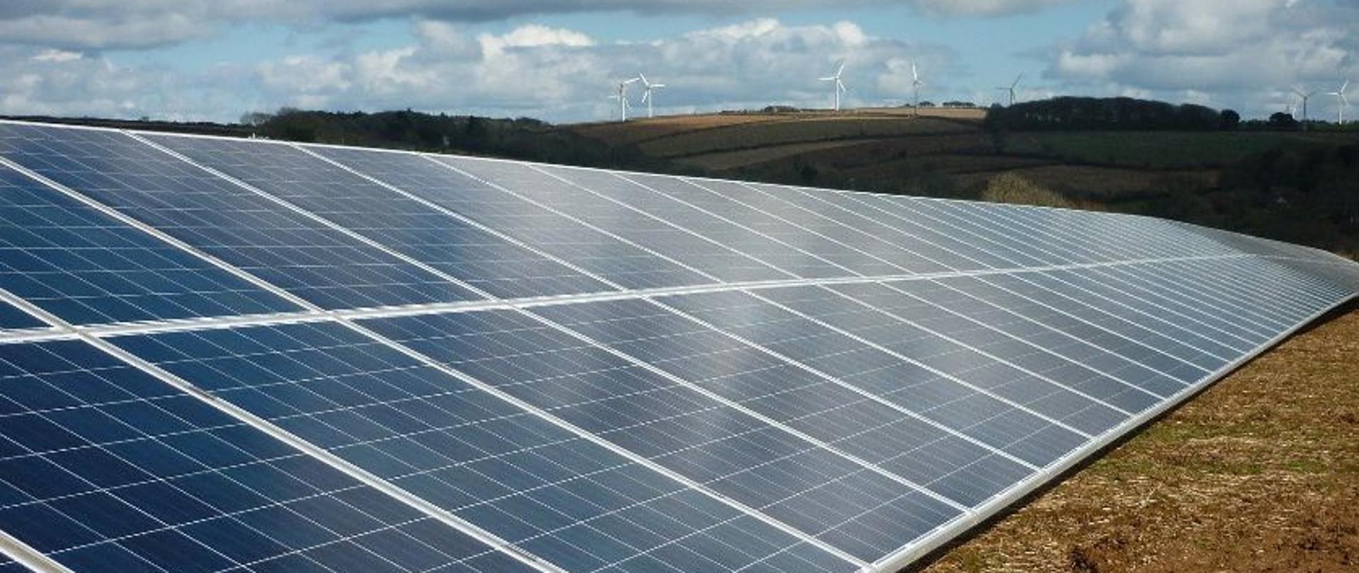 Na pierwszym polanie panele fotowoltaiczne, w tle krajobraz rolniczy i elektrownie wiatrowe.