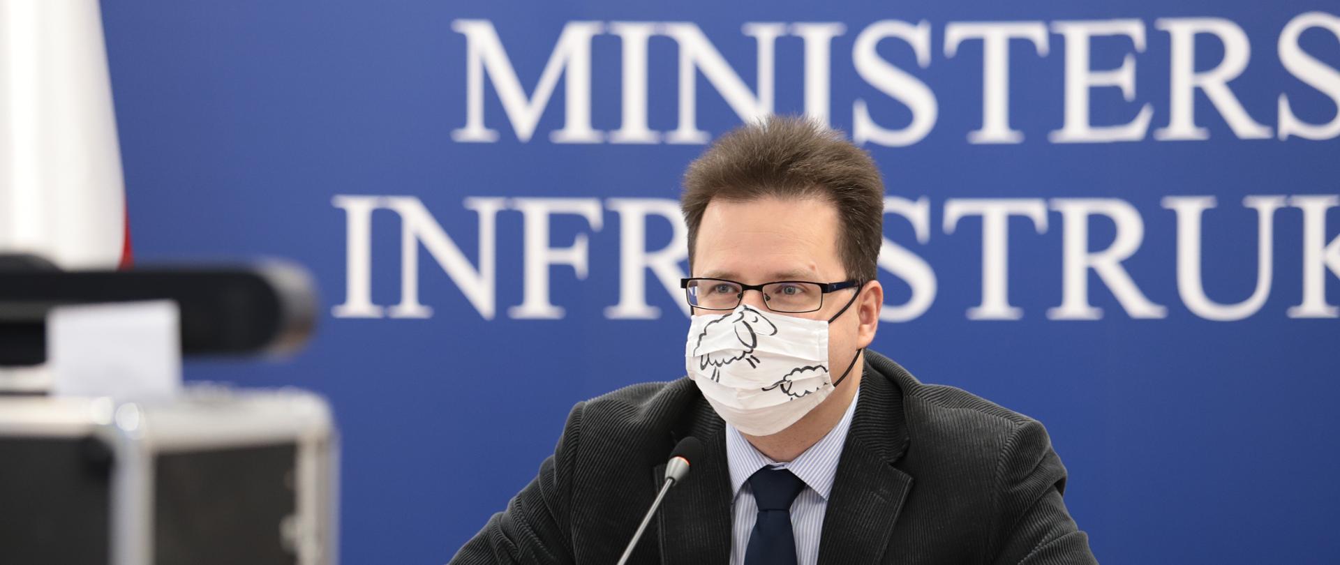 Na zdjęciu widać mężczyznę w ciemnej marynarce i jasnej koszuli. W tle stoi granatowa ścianka z napisem Ministerstwo Infrastruktury. Mężczyzna siedzi przy ciemnym stole, ma na twarzy białą maseczkę.