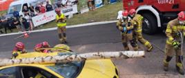 Strażacy podczas ćwiczenia ratowniczego. Żółty samochód osobowy przygnieciony powalonym drzewem. W tle banery reklamowe, samochody dostawcze oraz namioty strażackie. 
