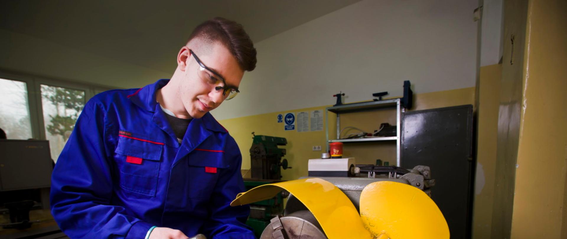 uczeń stoi przy tokarce, dokonuje pomiaru średnicy materiału