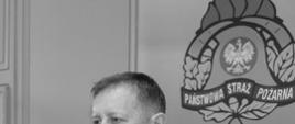 czarno białe zdjęcie przedstawiające Macieja Dobrakowskiego w mundurze wyjściowym za nim rollup komendy miejskiej PSP w Piotrkowie Trybunalskim