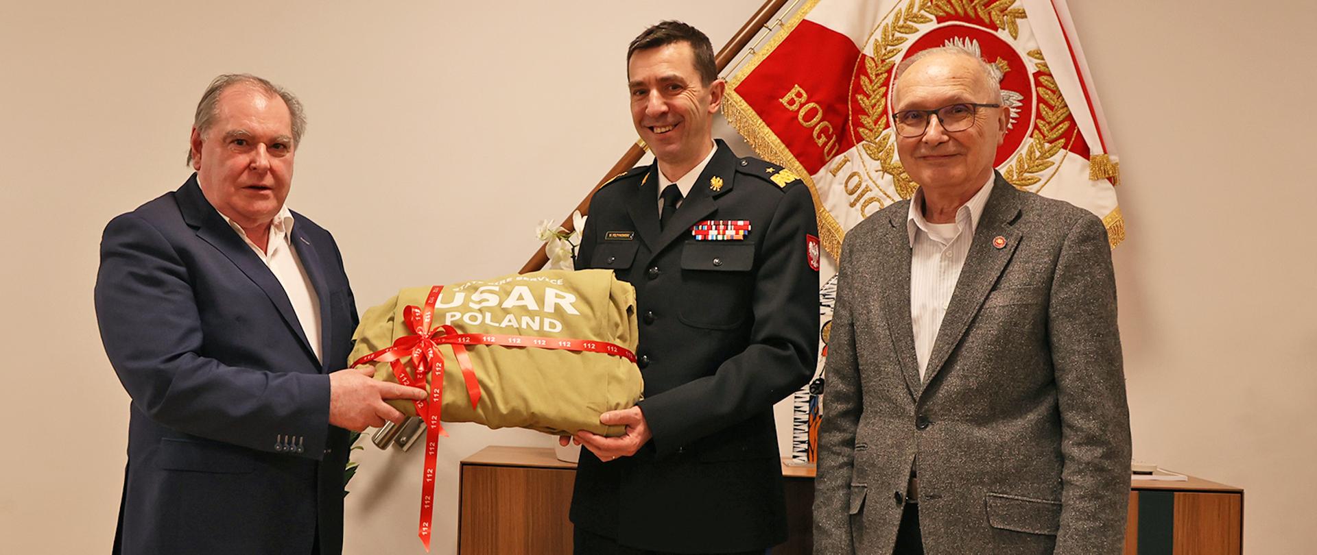 Dwaj mężczyźni w marynarkach oraz strażak w mundurze galowym w trakcie przekazania przedmiotu licytacji na tle sztandaru Komendy Głównej Państwowej Straży Pożarnej 