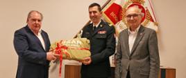 Dwaj mężczyźni w marynarkach oraz strażak w mundurze galowym w trakcie przekazania przedmiotu licytacji na tle sztandaru Komendy Głównej Państwowej Straży Pożarnej 
