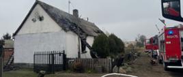 Na zdjęciu widać dom jednorodzinny, który prawdopodobnie uległ pożarowi. Dach budynku jest nadpalony i częściowo zniszczony, a na miejscu znajdują się wozy strażackie oraz strażacy prowadzący akcję gaśniczą. Na ziemi rozłożone są węże strażackie, a w pobliżu ogrodzenia leży sprzęt gaśniczy. Pogoda jest pochmurna, a wokół domu widać drogę, płot i roślinność.