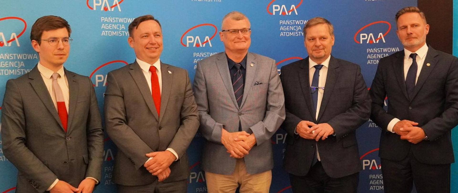 Na zdjęciu widzimy pięciu mężczyzn stojących na tle ścianki z logotypem Państwowej Agencji Atomistyki (PAA) – polskiej instytucji zajmującej się nadzorem nad bezpieczeństwem jądrowym i ochroną radiologiczną.
Wszyscy mężczyźni są elegancko ubrani w garnitury i krawaty, co sugeruje oficjalne wydarzenie, takie jak konferencja prasowa, spotkanie robocze lub podpisanie umowy. Tło zdjęcia to niebieska ścianka z logo PAA i napisem „Państwowa Agencja Atomistyki”, które pojawia się wielokrotnie.
Mężczyzna pośrodku (trzeci od lewej) wyróżnia się jasnoszarą marynarką i okularami – możliwe, że pełni on ważną rolę w agencji. Pozostali panowie są również ubrani w formalne stroje, a niektórzy z nich mają przypięte znaczki, co może świadczyć o ich stanowiskach lub reprezentowanych instytucjach.
Zdjęcie ma oficjalny, profesjonalny charakter.