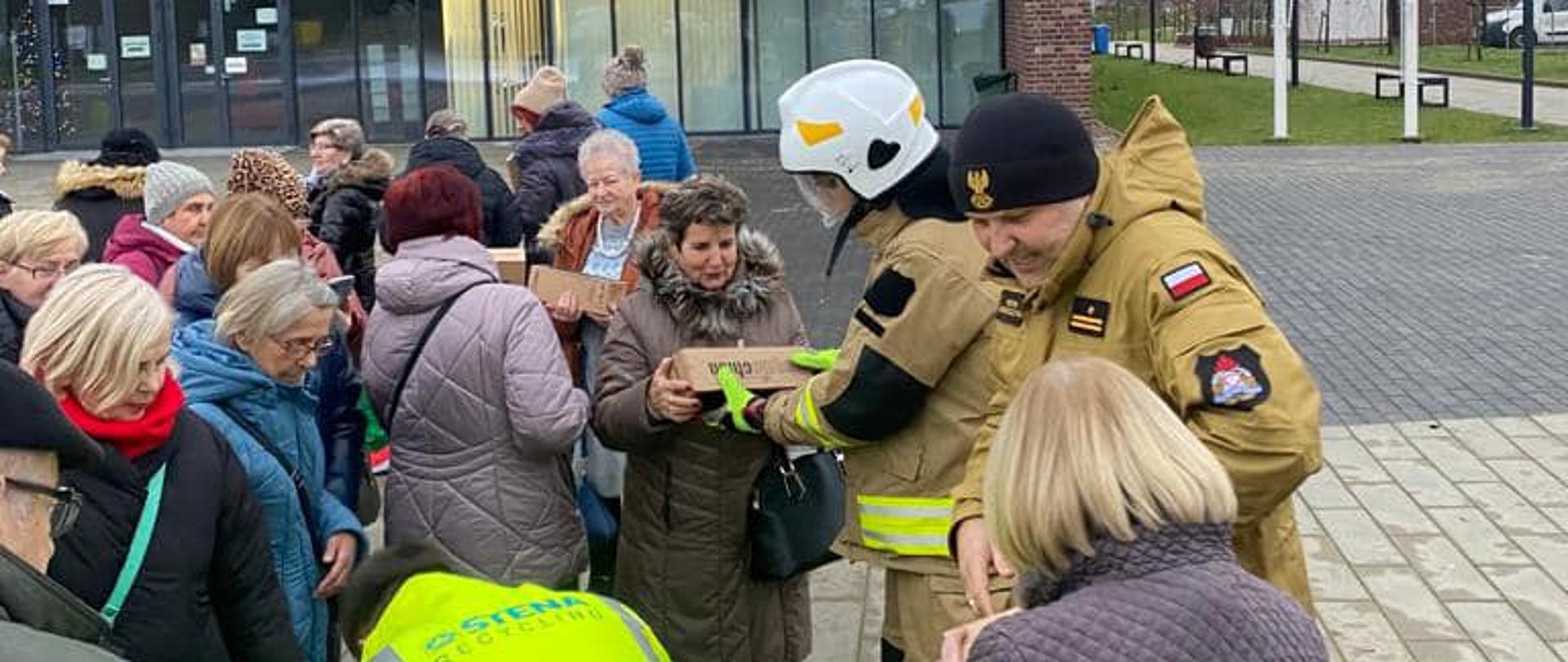 Na zdjęciu brukowany plac, na którym znajduje się w swobodnym ustawieniu skupisko dużej grupy osób ubranych w płaszcze i kurtki, część w nakryciu głowy. Wśród zgromadzonych widoczne dwie osoby, jedna z nich w białym strażackim hełmie, druga w czarnej czapce z orzełkiem, w piaskowych kurtkach mundurowych z emblematami na rękawach, w tym białoczerwoną naszywką. Strażak w hełmie przekazuje jednej z osób prostokątny kartonik. W tle fragment nowoczesnej, przeszklonej elewacji budynku. Niebo jasne, całkowicie zachmurzone.