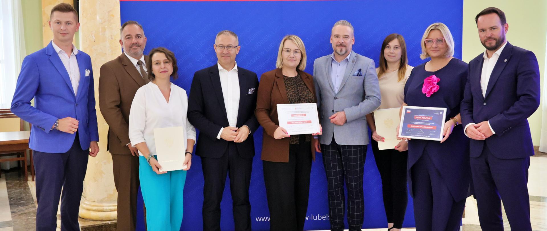 Duża grupa dziewięciu osób – sześciu mężczyzn i trzy kobiety – stojących przed niebieską ścianką z napisem 'Lubelski Urząd Wojewódzki w Lublinie'. Kilku z nich trzyma dokumenty lub certyfikaty.