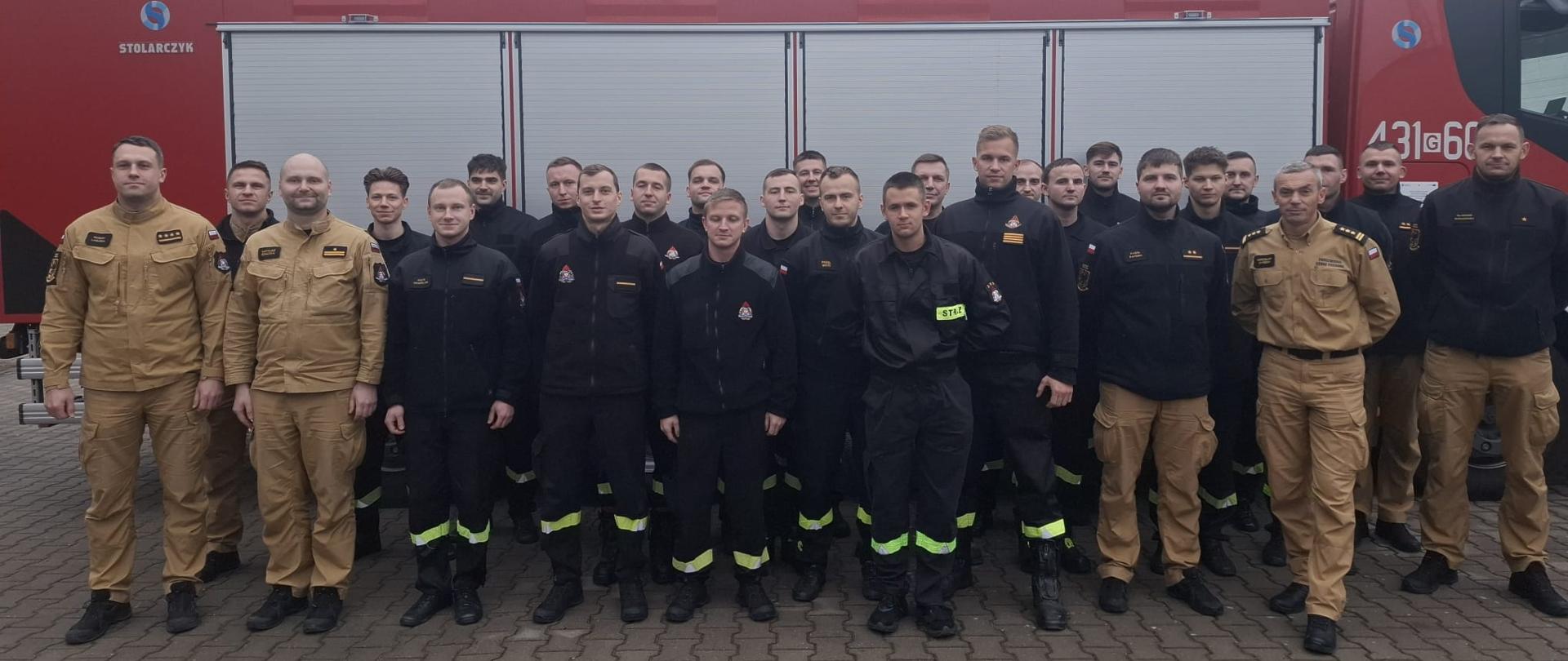 Parunastu strażaków pozujących do zdjęcia z okazji ukończenia kursu. Funkcjonariusze ubrani w ubrania koszarowe stoją na kostce brukowej. W tle samochód straży pożarnej.
