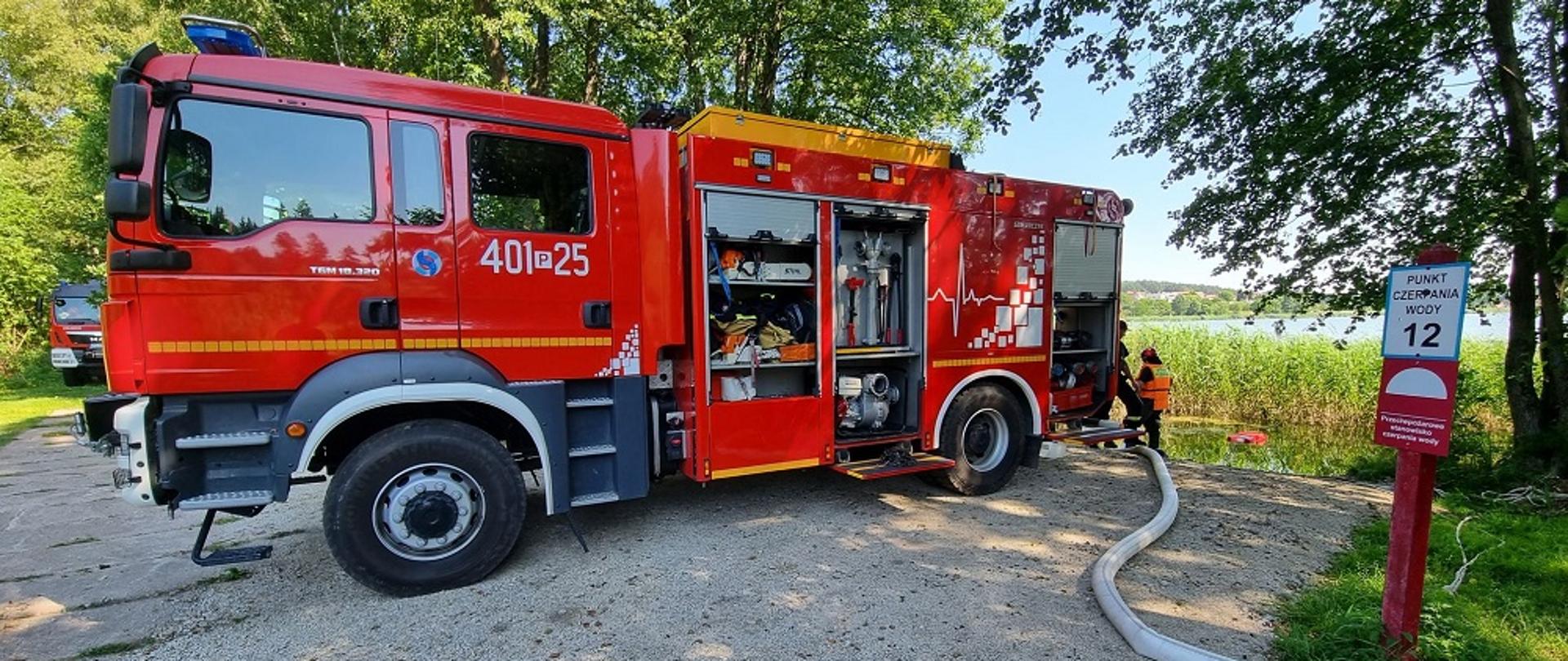 Zdjęcie przedstawia strażaków oraz samochód gaśniczy podczas ćwiczeń nad jeziorem z pompami w ramach doskonalenia zawodowego.
W tle jezioro i las. 