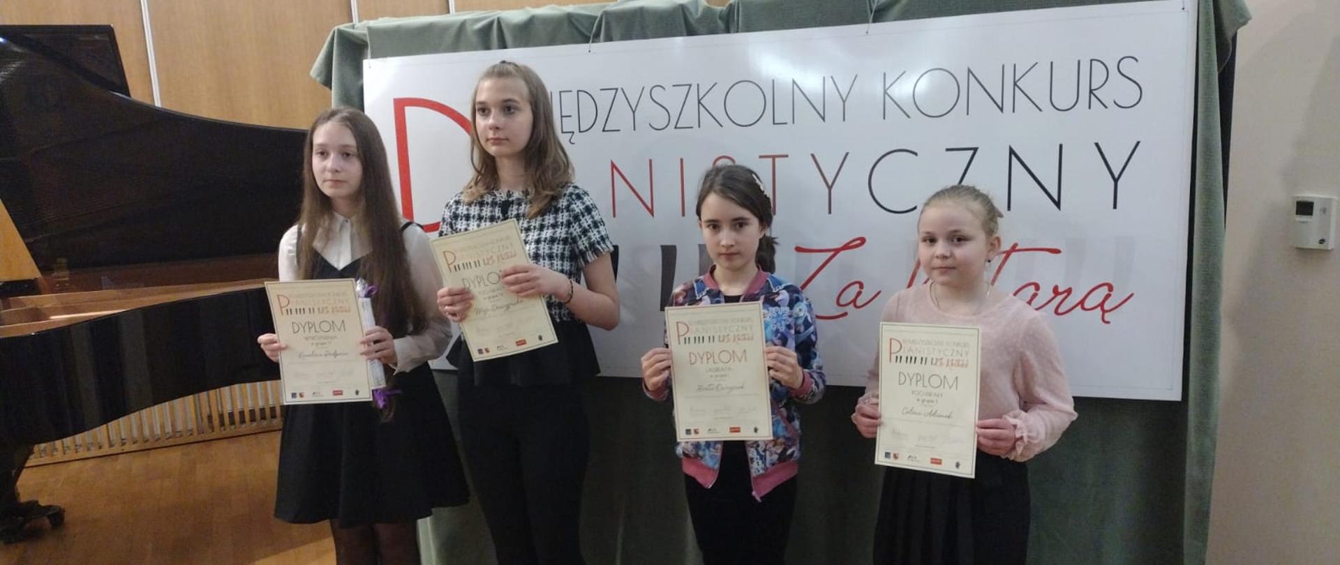 Zdjęcie przedstawia uczennice klasy fortepianu naszej szkoły na tle baneru konkursowego. Uczennice trzymają w rękach zdobyte dyplomy.