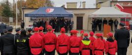 Druhowie zaproszeni goście oraz młodzieżowa drużyna pożarnicza stoją na uroczystości przed remizą.