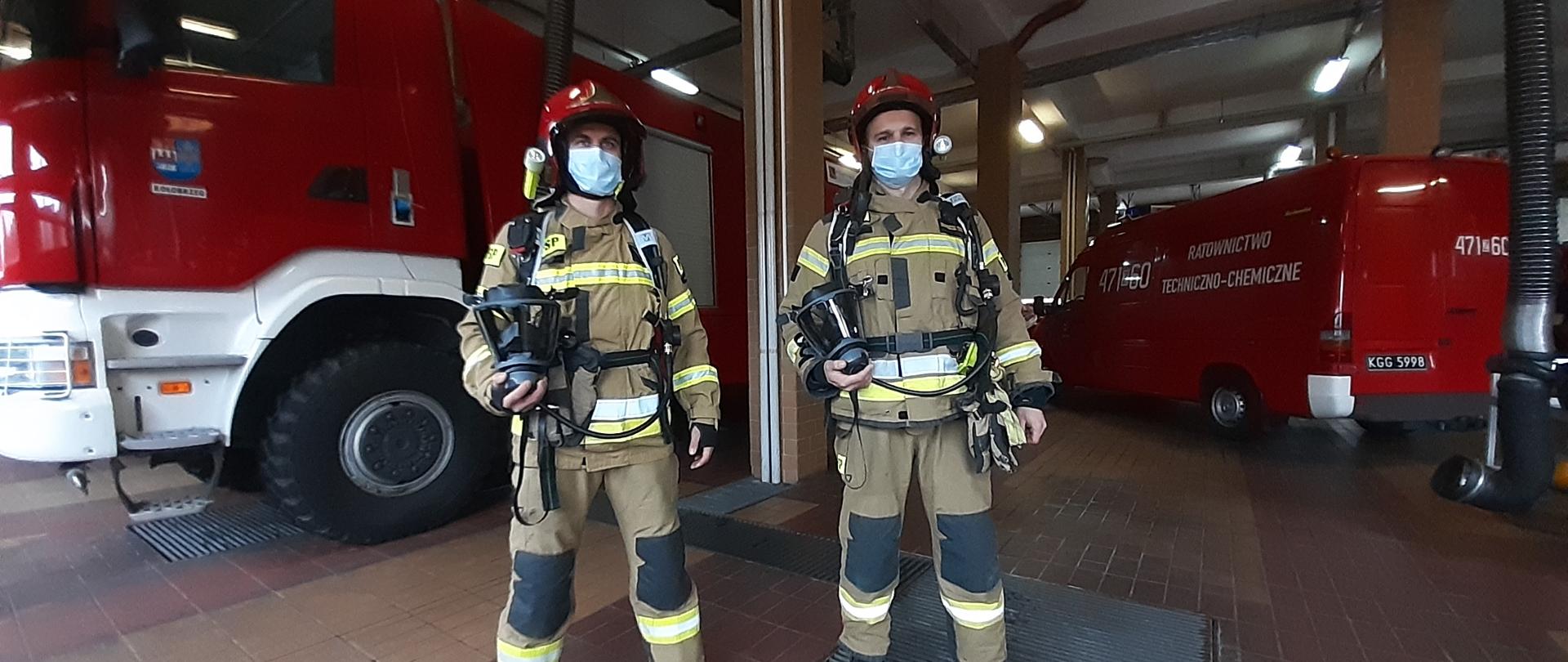 Dwóch strażaków ubranych w sprzęt ochrony dróg oddechowych na tle samochodów pożarniczych