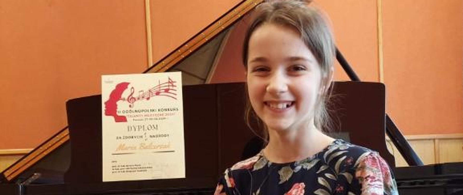 Zdjęcie kolorowe. Uśmiechnięta dziewczynka w sukience w kwiaty siedzi przed otwartym czarnym fortepianem. Na pulpicie fortepianu stoi dyplom z konkursu.