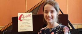 Zdjęcie kolorowe. Uśmiechnięta dziewczynka w sukience w kwiaty siedzi przed otwartym czarnym fortepianem. Na pulpicie fortepianu stoi dyplom z konkursu.