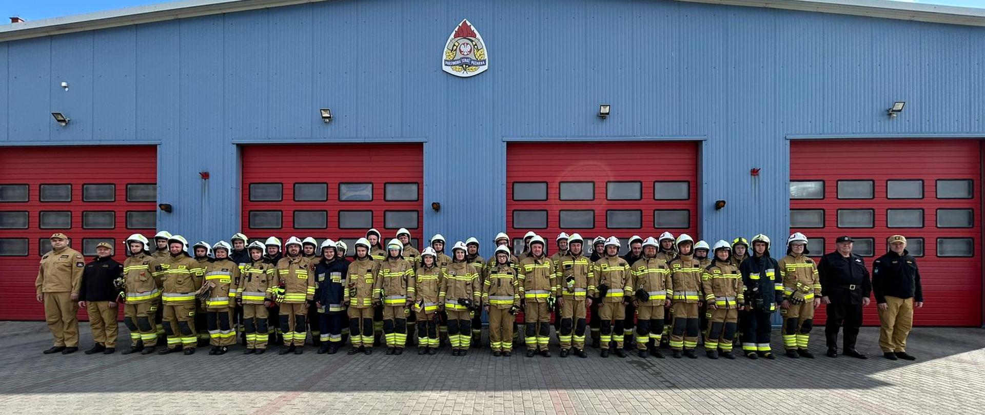 Na zdjęciu strażacy ochotniczych straży pożarnych na zakończeniu szkolenia podstawowego. Strażacy w ubraniach specjalnych znajdują się przed garażem JRG Jędrzejów
