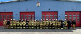 Na zdjęciu strażacy ochotniczych straży pożarnych na zakończeniu szkolenia podstawowego. Strażacy w ubraniach specjalnych znajdują się przed garażem JRG Jędrzejów