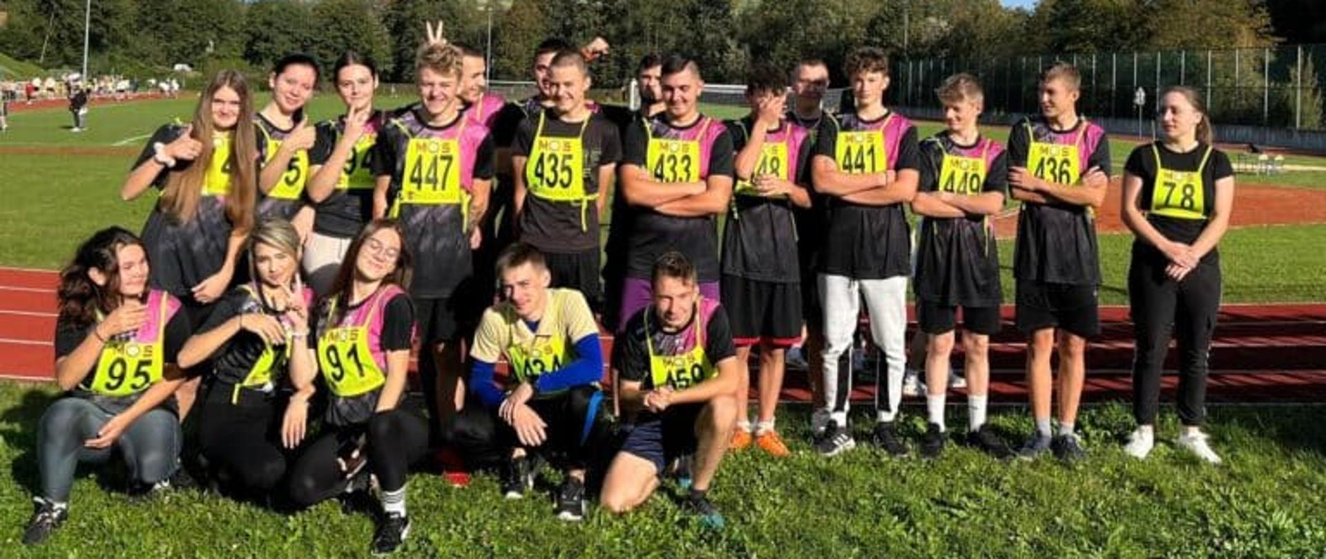 Pamiątkowe zdjęcie grupowe młodych sportowców w czarno-różowych strojach z numerami startowymi „MOS”. Część osób stoi, a część kuca na trawie obok czerwonej bieżni lekkoatletycznej w słoneczny dzień.